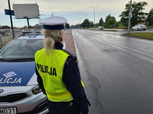 policjantka przy radiowozie obserwuje ruch pojazdów na drodze o dwóch pasach ruchu w jednym kierunku